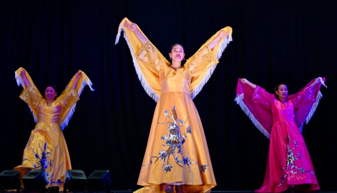 Three Filipino women in colourful dresses lift up their arms during a Filipino dance presentation at Carassauga Festival