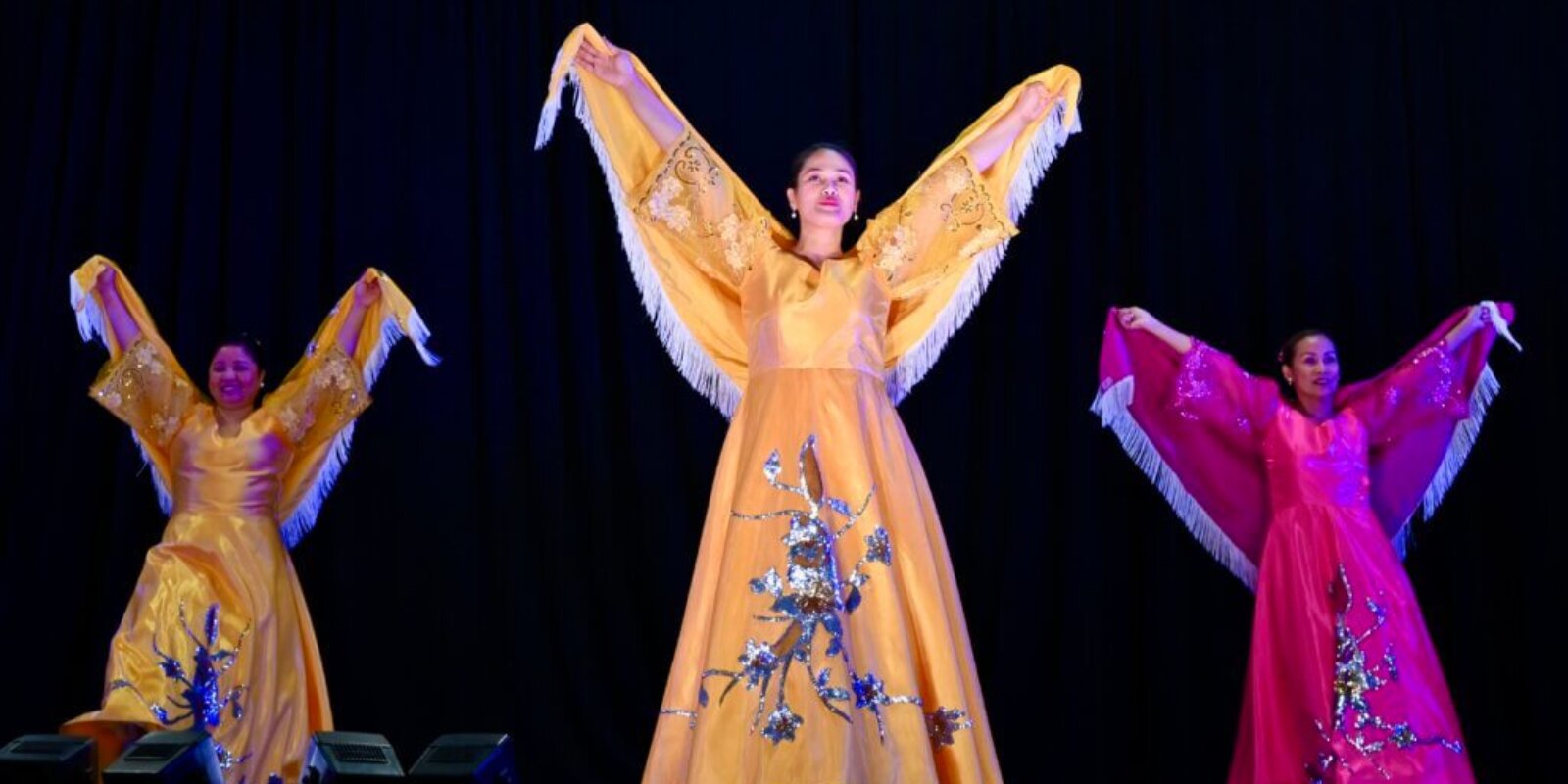 Three Filipino women in colourful dresses lift up their arms during a Filipino dance presentation at Carassauga Festival
