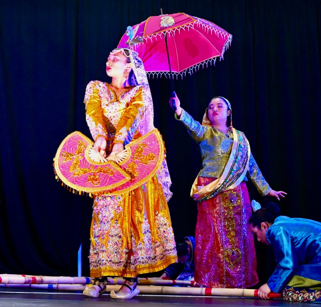 Two Filipino female dancers are shown, with one holding two pink fans and the other holding a pink umbrella. A third person is holding two bamboo poles on the ground.