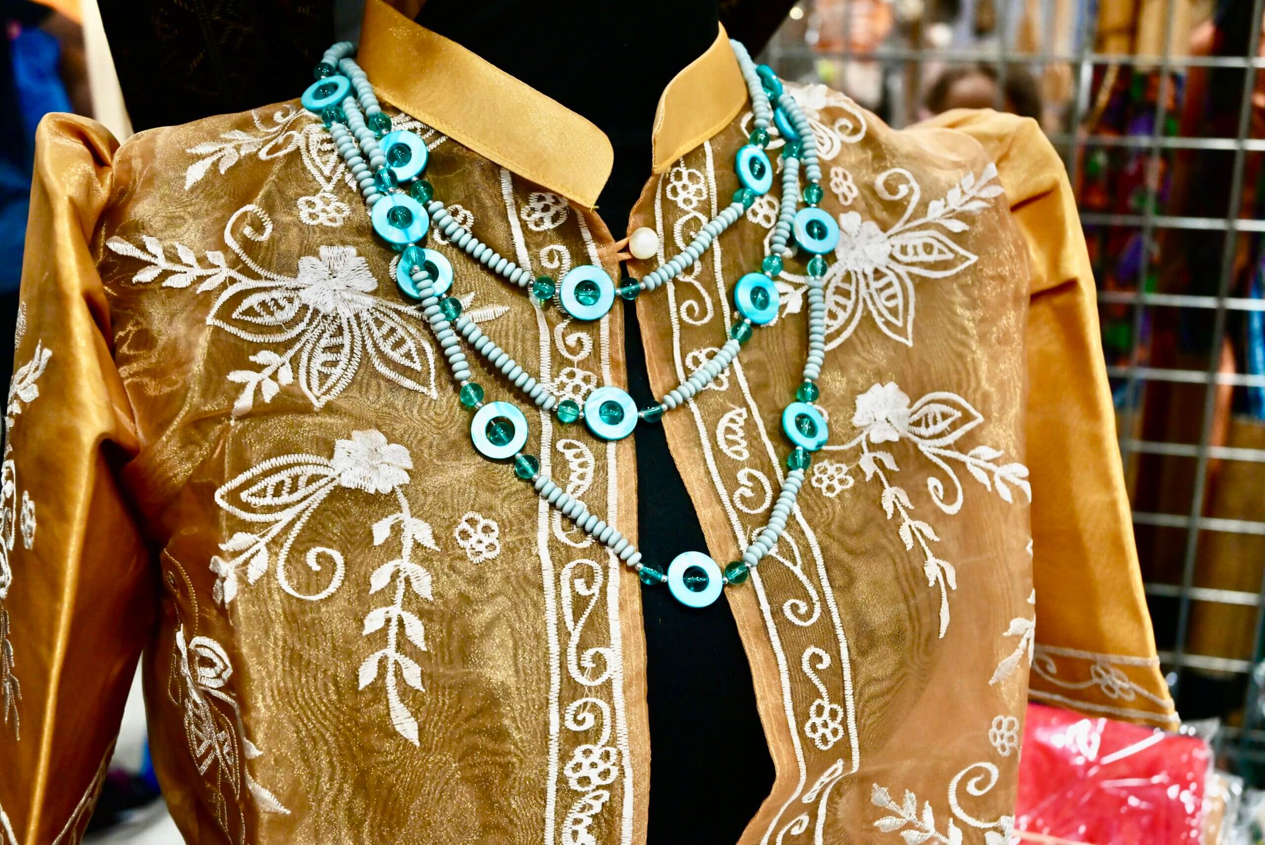 A picture of a gold, see-through, bolero style formal dress shirt with intricate white embroidery is shown with a blue three-chained beaded necklace.