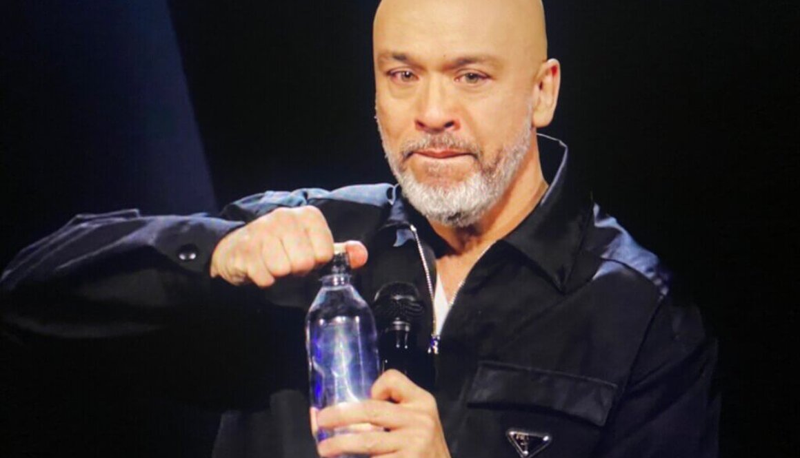Filipino comedian Jo Koy is shown opening a water bottle and holding a microphone. His lips are pursed, his eyes are misty with tears.
