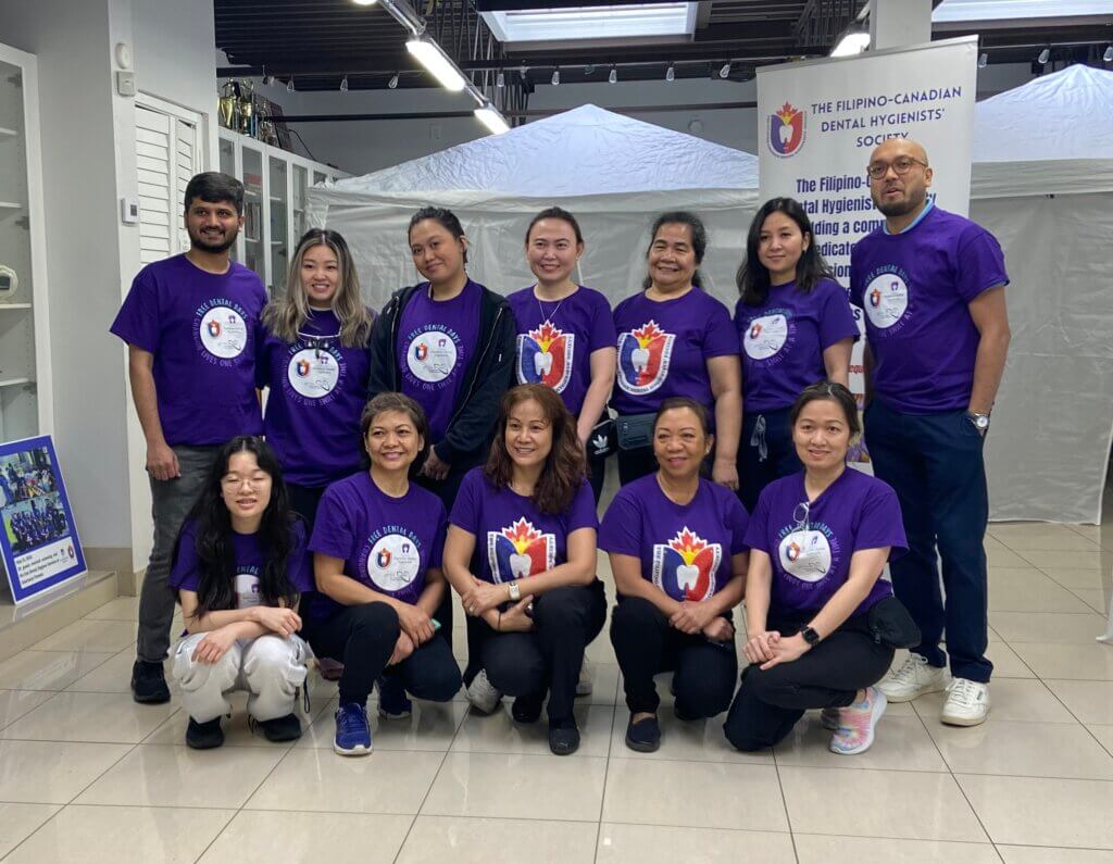A dozen members of the Filipino-Canadian Dental Hygienists' Society pose for a photo in a hallway with a banner in the background.
