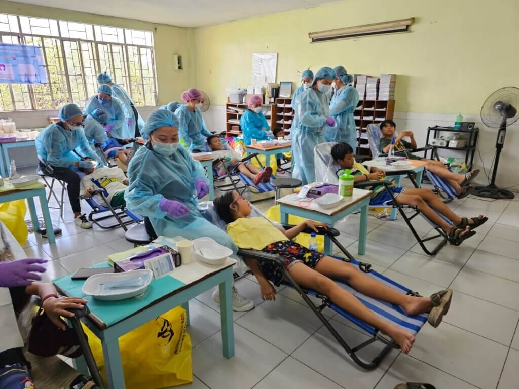 Children sit on camping chairs as dentists and hygienists in blue medical gowns do dental work.