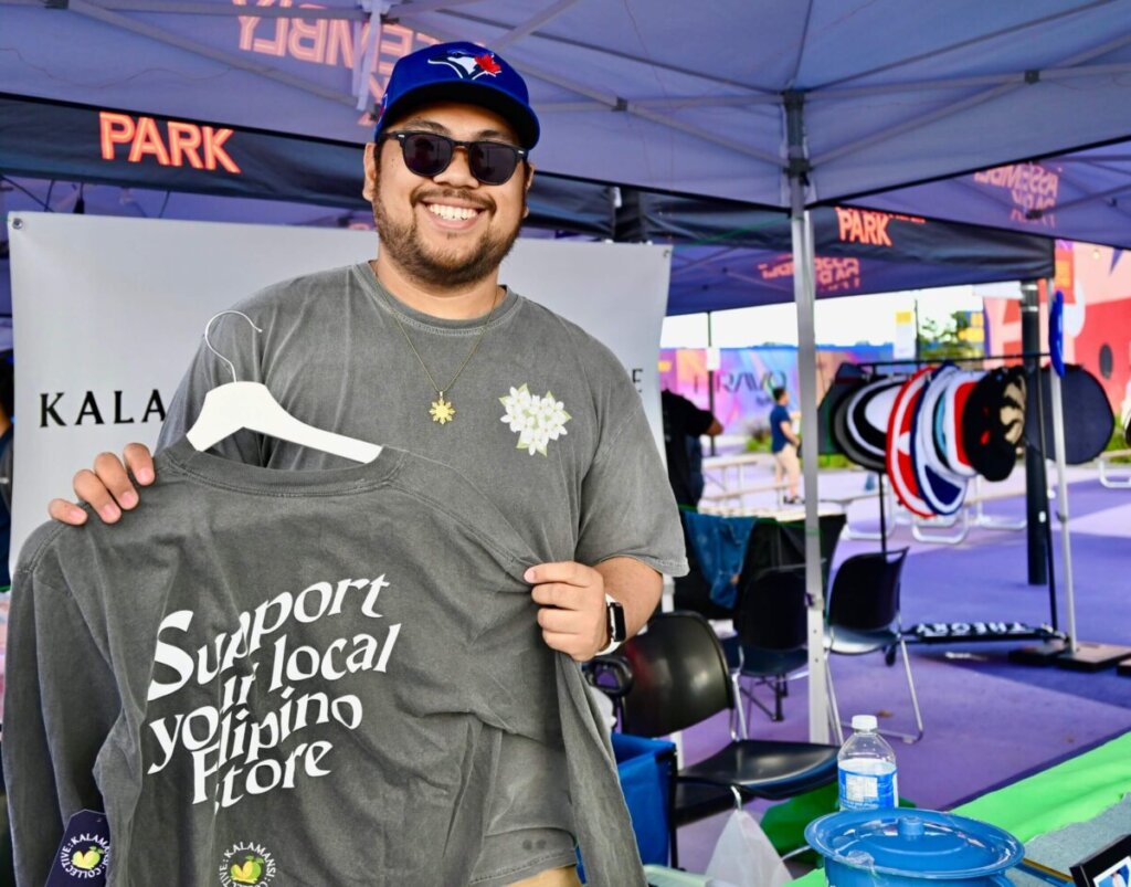 Kalamansi Collective founder Kris Pangilinan is shown at his booth at a market, holding up a gray shirt with the words "Support your local Filipino Store"