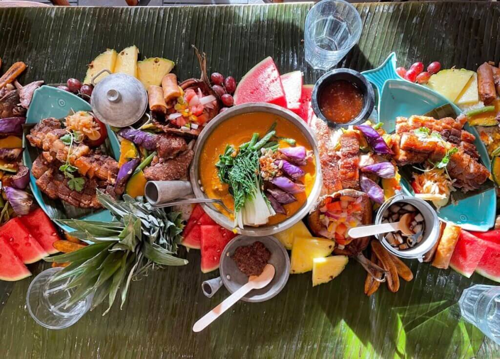 Banana leaves are spread out on a table with a full spread of fruits, vegetables and a variety of Filipino dishes.
