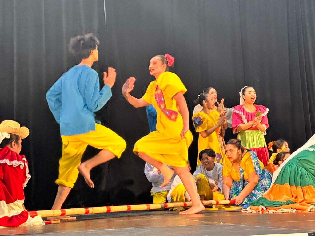 Filipino dancers in colourful attire dance the tinikling, as they hop and jump in and out of two clapping bamboo poles.